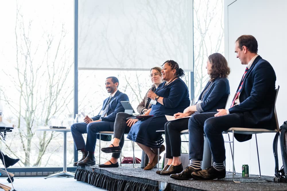 Panelist speaking into a microphone while fellow speakers listen on a conference stage