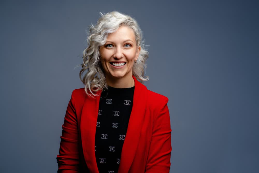 Smiling professional with silver curls wearing a red blazer against a dark blue studio background