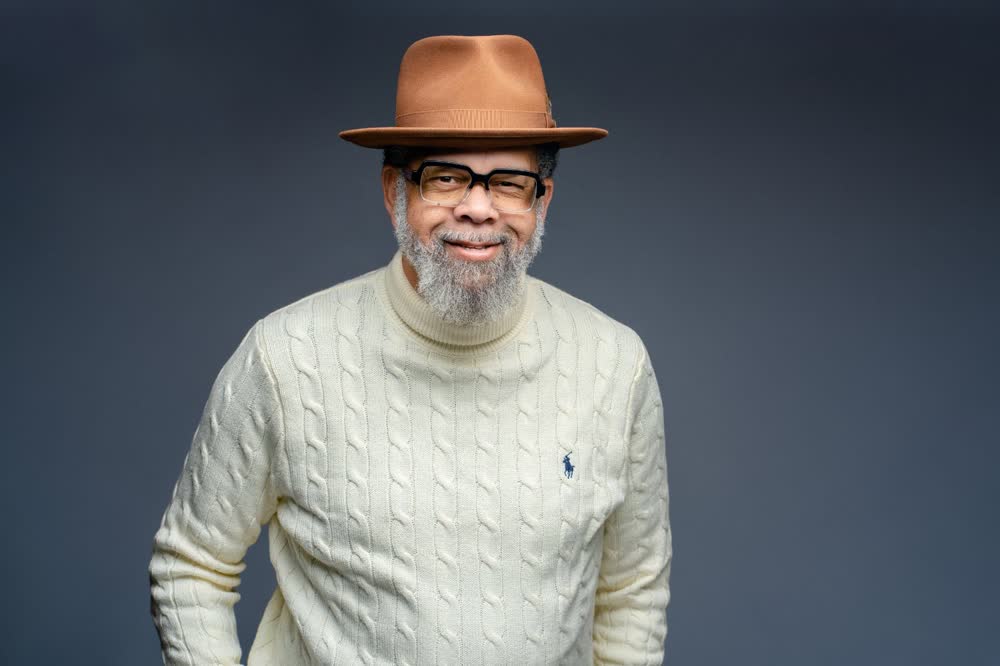 Smiling professional in glasses, a brown hat, and a cream cable-knit sweater against a dark backdrop