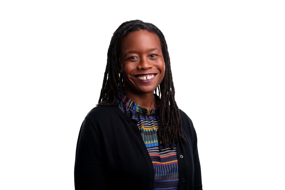 Smiling professional with long locs wearing a patterned blouse and black cardigan on a white background