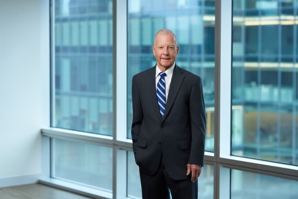 Executive standing in a suit beside large office windows overlooking nearby buildings