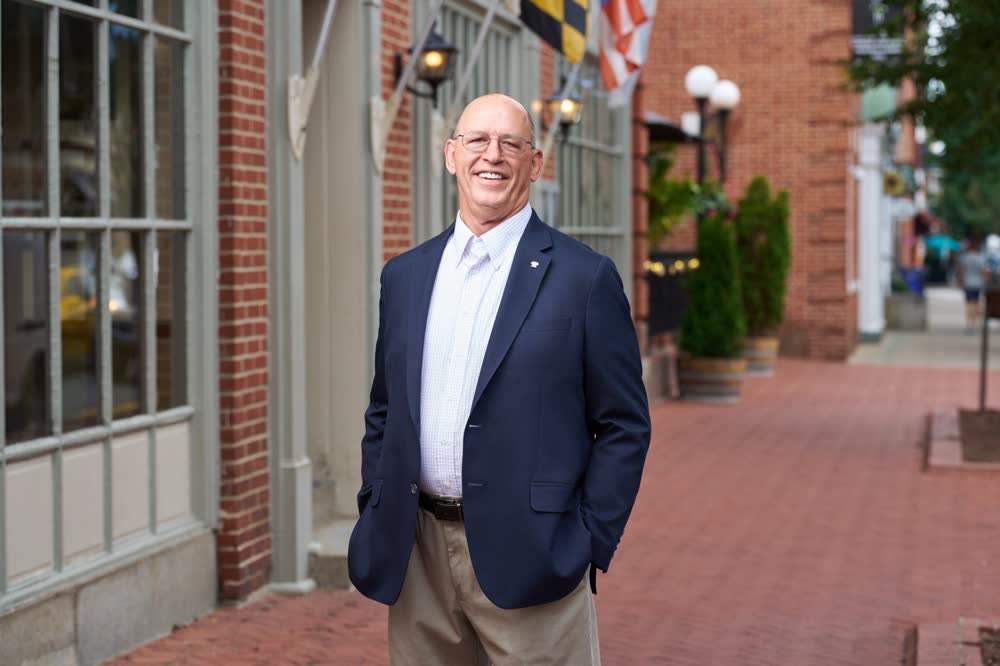 A business professional smiling with both hands in his pockets on a brick sidewalk